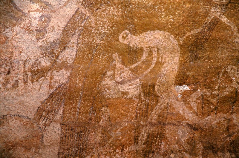 Oued Djaret, Algeria. Woman with elongated body bends left holding possible insect and bag in outstretched hands.  Image ID: algdja0080065