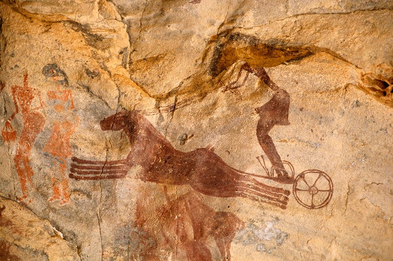 Tassili n'Ajjer, SE Algeria. Maroon painting of two horses galloping to left drawing chariot with charioteer wearing skirt and holding four reins. Image ID: algtdj0010009