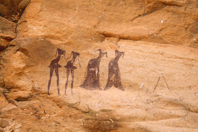 Tarssed Jebest, Algeria. Line of four maroon figures face forwards. Two men at left with very thin spike heads short skirts Two women at right with very thin spike heads. Image ID: algtdj0010030
