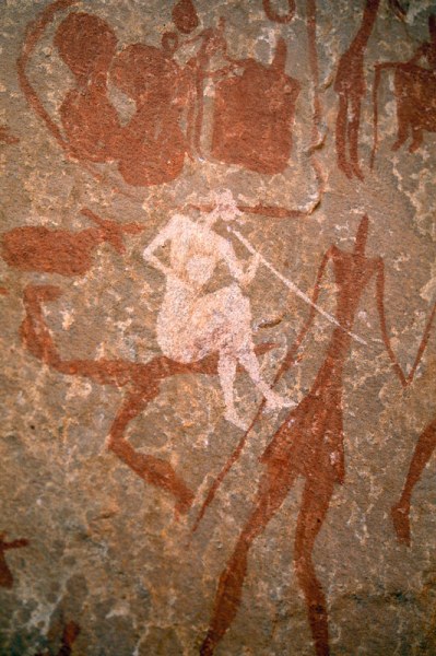 Tadrart, Algeria. White seated man with legs crossed and wearing cap, clothes and pointed shoes holds stick (musical instrument?) to mouth. Image ID: algtdr0030006