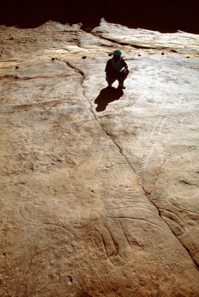Oued Djerat, Algeria. Tuareg crouches by shoulder of huge engraved giraffe. Giraffe’s head faces right (left in photograph) in foreground. Image ID: algtod0140006