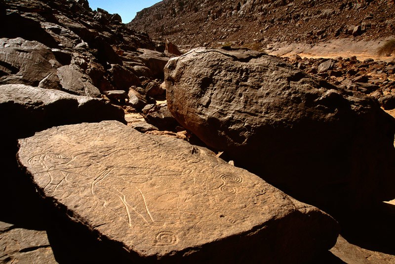 Oued Djerat, Algeria. Oblique view of deeply incised man facing right. He wears helmet, back skirt and bracelets, has huge testes hanging between parted legs and holds loaded bow in one hand. Image ID: algtod0180011