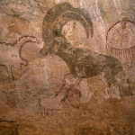 Djanet, Algeria. Close-up of red Barbary sheep, oval with dependent rays and schematic cow(?) outlined in red. Note red outlined animal between sheep’s legs and torso of person with penis behind sheep’s horn. Image ID: algdja0030014