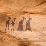 Tarssed Jebest, Algeria. Line of four maroon figures face forwards. Two men at left with very thin spike heads short skirts Two women at right with very thin spike heads. Image ID: algtdj0010030
