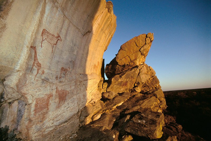 Tsodilo Hills. Finger paintings in red of eland and giraffe, and positive handprints, with setting sun lighting right-hand side. Image ID: bottsd0040004