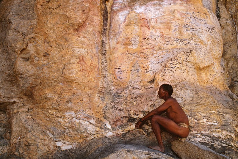 Tsodilo Hills. Above steep rock slope to right of White Paintings Shelter, Zhu man seated before panel of eroded animal paintings that includes antelope facing left and striped animal. Image ID: bottsd0620001