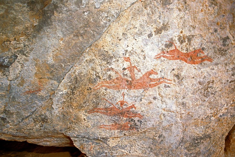 Archei Guelta. Outside cave on exposed sandstone cliff. Four red horses, two with armed riders, gallop right. At left, horizontal human figure flies after horses. Horse Period. Image ID: chaarg0020004