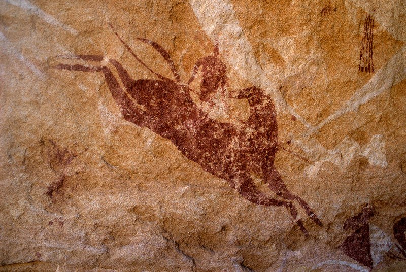 Ennedi Plateau. White ‘running’ shapes superimposed by red leaping horse with head turned back mounted by red man holding long spear. Bottom right, bichrome shield. Top right, red camel-saddle trappings. Image ID: chaenp0050015