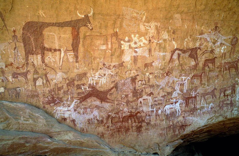 Ennedi Plateau. Huge panel of mainly bichrome paintings on rear wall of cave. Top left, large bichrome cow with udder facing right superimposing outline white cow facing left. Bichrome man stands behind large cow holding arrow(?). Numerous cattle, woman in skirt, running red camel, goats and sheep surround big cow. To right of cow, white geometric shapes are remains of bichrome cattle. Below, red and white horses with horsemen holding swords. Numerous small people facing forwards. Image ID: chaenp0050021