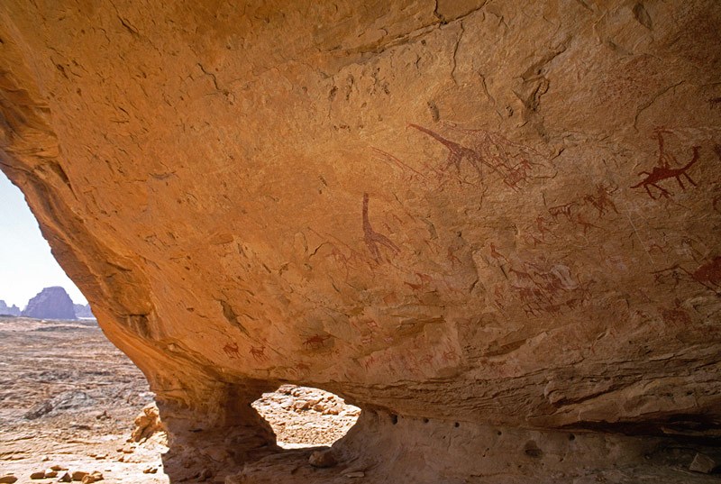 Tassili d' Emi Koussi. Looking out from the entrance of a large sandstone cave on the lower slopes of Emi-Koussi. The floor slopes up to the back wall for some 30 metres, rapidly narrowing towards the back. The lefty wall is profusely painted, many images being in polychrome, with 200 and more images of people, armed warriors, cattle, camels, giraffe, antelope, an elephant and human figure touching, people surrounding what may be a wild cow, and geometric designs. Most paintings belong to the Camel Period. The cave is still used by nomadic groups as a resting place. Image ID: chatek0070002