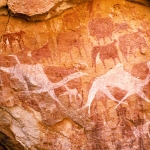 Ennedi plateau. Wall of shallow sandstone cave. Bichrome images of cattle with white udders and person with round head, facing forward and flanked by two small figures, are all superimposed by white camel running to right. Horse and Camel Periods. Image ID: chaenp0020022