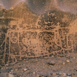Tibesti Mountains. Recorder drawing pecked engravings on wall of gorge. Crude engraving of large cow in background. Note graffiti in Roman (HASSN) and Arabic script and dates. Image ID: chatim0010015