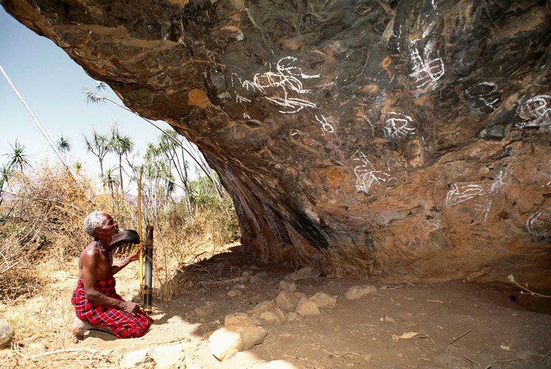 Laikipia. Ndorobo man with bow and quiver of arrows kneels at a rock shelter adorned with white symbolic paintings suggesting meat-feasting rituals.  Image ID: kenlai0060004