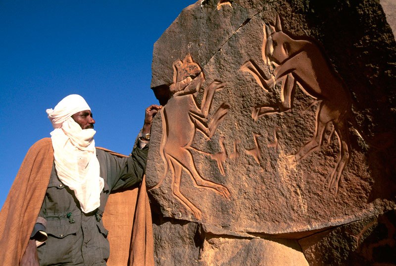 Messak. Tuareg guide standing beside so-called panel of “fighting cats”. Image ID: libmes0040128