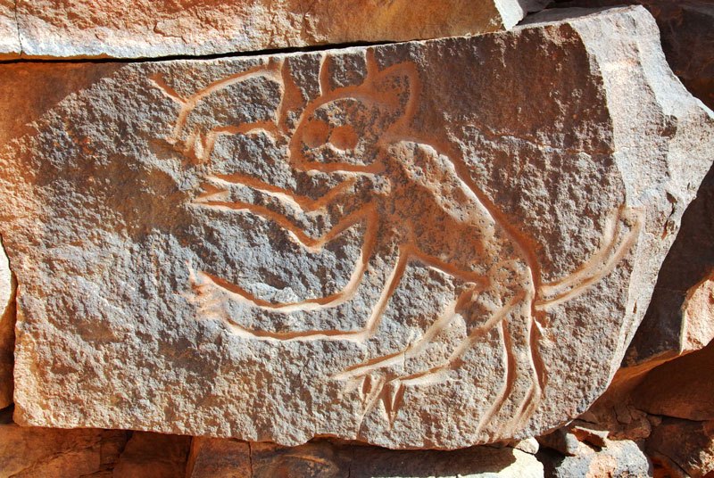 Messak. Details of engraved predator with two large eyes, spotted body and claws standing, facing left, arms outstretched with small animal (cow?) by its face. Probably early hunter period. Image ID: libmes0170017