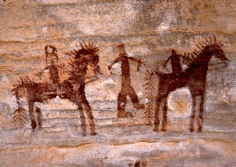 Tagant, Mauritania. Two dark maroon men, one wearing headdress, riding horses with fancy manes and tails separated by man on foot. Image ID: mauaio0010001