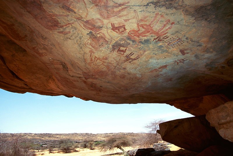 Tagant, Mauritania. On bulge between back wall and roof of shelter, thickset red man facing forwards, red cows, red cow with elongated legs, red shape (predator?), red tridents on vertical crossed bars. Image ID: mautag0080013