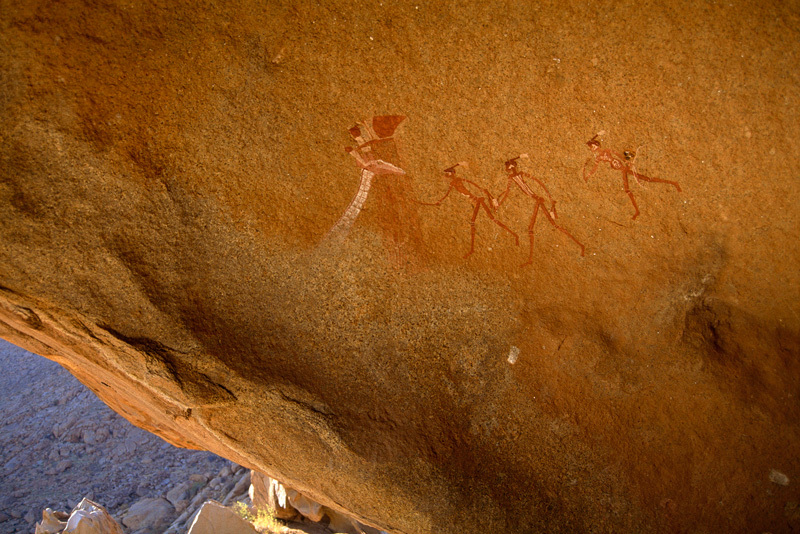Polychrome Bushman/San paintings apparently depicting the head and white neck of a giraffe facing right approached by 3 decorated figures (on right). The paintings are located behind a narrow ledge high up on the Brandberg massif and it is probable that this was ritual site, perhaps a rain making site. Image ID: nambrg0030025