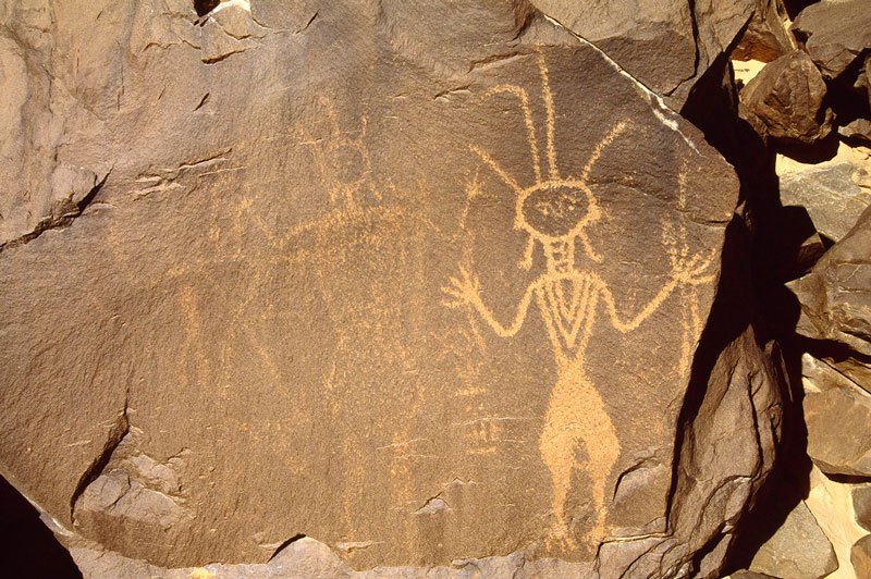 Air Mountain, Niger. Faded Libyan Warrior. Libyan Warrior with round head and face, wearing four feathers and dangling earrings on head, beeches and decorated vest, hands raised one holding two javelins with metal heads. Image ID: nigeam0010081