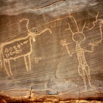 Western Air Mountains, Niger. Pecked outline domestic bull with body decorated with two horizontal lines of ancient Tifinagh script. To right of bull, slim Libyan warrior with round outline head and three feathers, long neck and one earring, striped jacket and breeches, raised hands one holding elongated leg of schematic ostrich. Image ID: nigwam0050024