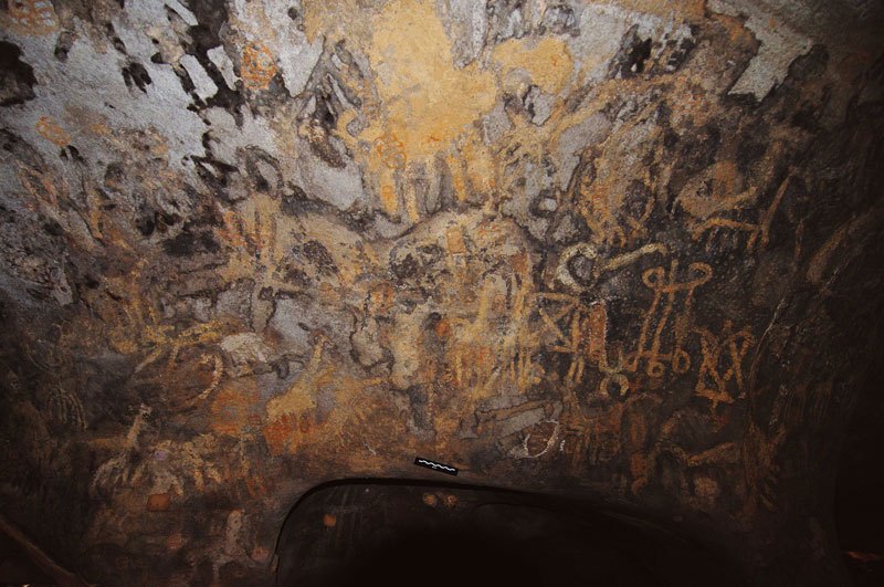 Dhayamole. Body and legs of orange, five-legged giraffe beside black figure(?) walking right. Image ID: somdha0020017