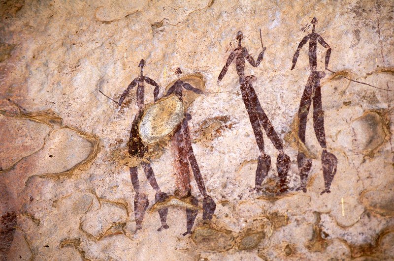 Four tall men. Four red naked men face forwards holding sticks. Two men have emissions from faces and knee bands. Image ID: soaefs0010023