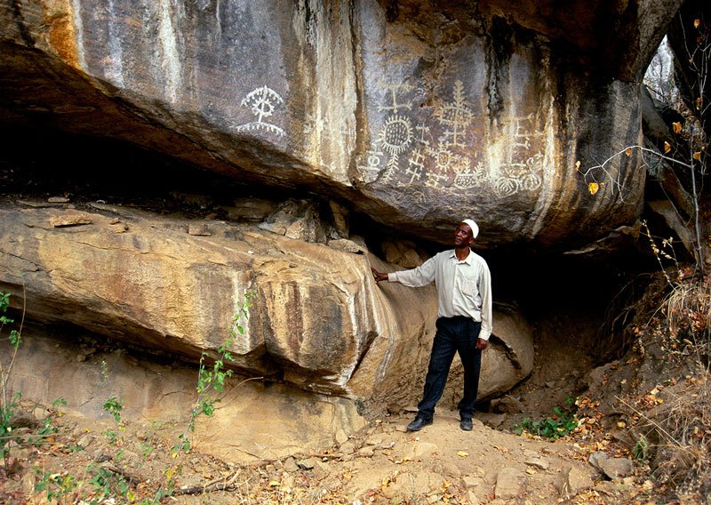 Overhanging granite boulders with protected area. Site has been vandalised by entrepreneurs digging for gold believed buried here by Germans during 1914-1918 War. Official custodian stands below large panel of Late White geometric designs. Image ID: tankon0020043