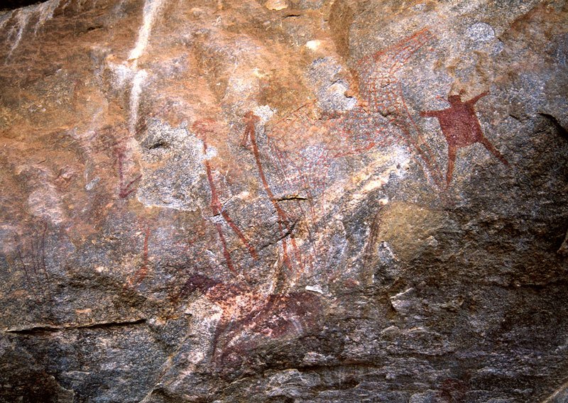 Large panel with many feint images of giraffe superimposed by red people and anthropomorphic figure. Line of four red outline decorated giraffes face right. Solid red anthropomorphic figure with animal ears and limbs outspread appears to ‘float’ in space. Image ID: tankon0030046