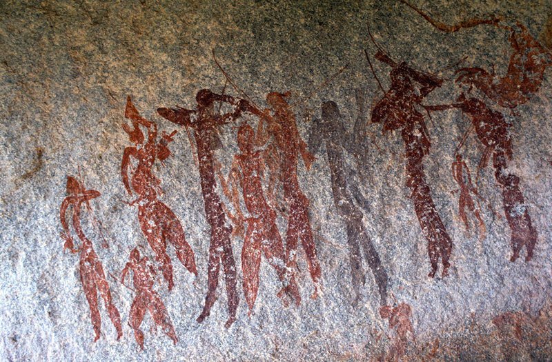 Mashonaland, Zimbabwe. Line of ten men. From left, red man with slender body, and inverted heart-shaped head holds bow and arrows in raised hand and plant(?) in other hand. Red man facing forwards wearing mask/headdress holds bow in one hand. Image ID: zimmsl0170006